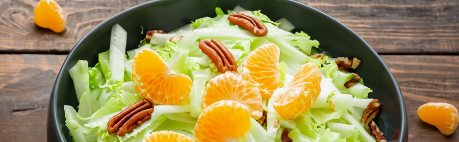 Een frisse salade met knapperige groene bladeren, sappige mandarijnpartjes en knapperige pecannoten, geserveerd in een donkergroene kom op een houten tafel.